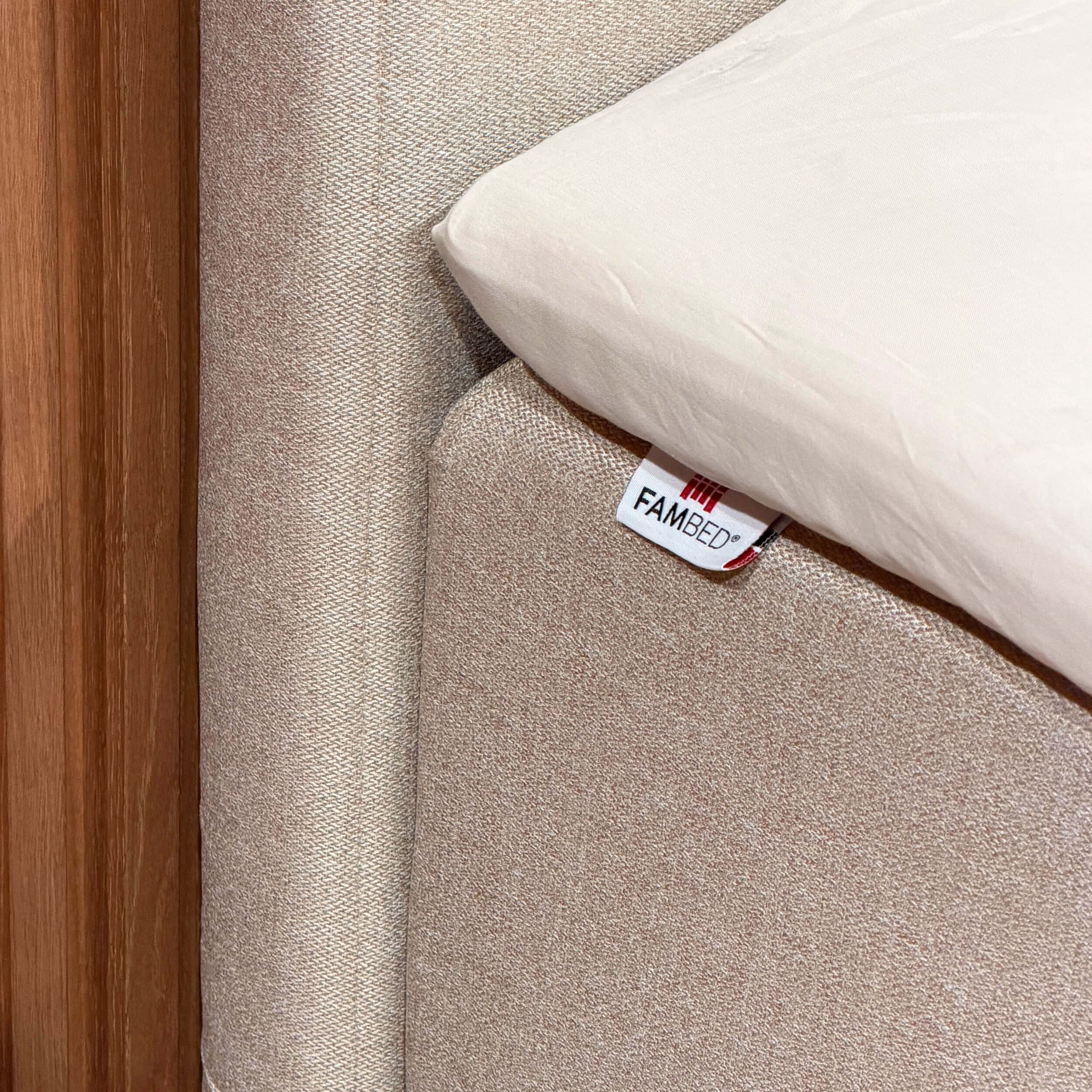 Primer plano de la funda beige que envuelve la esquina de la cama, con la etiqueta FAMBED y una sábana blanca encima