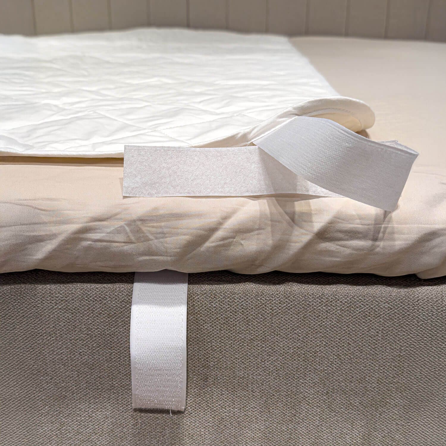 Esquina del protector de colchón blanco acolchado doblada hacia atrás para mostrar la correa de velcro que rodea el borde de la cama
