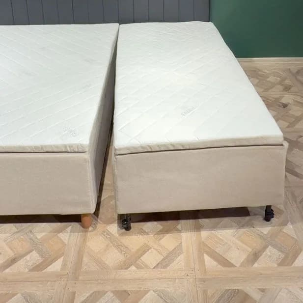 Dos módulos de cama completamente montados, colocados uno junto al otro sobre un suelo de madera clara y separados por un pequeño espacio. Cada módulo tiene su propia funda beige, su propio colchón acolchado y sus propias patas.
