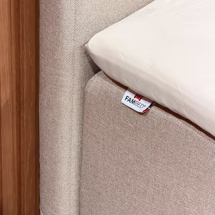 Close-up of the beige cover fitted around the corner of the bed, with a FAMBED label and white sheet on top
