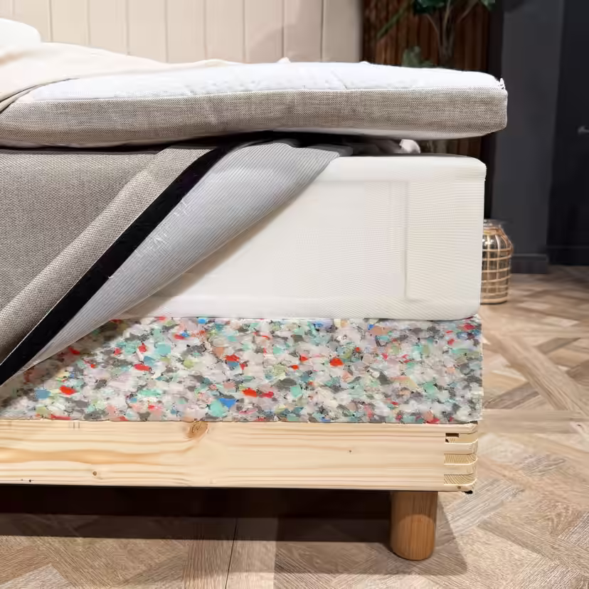 Bed layers opened at the corner, showing the mattress topper, sprung mattress with its cover opened and the bed base's foam edge above the wooden frame
